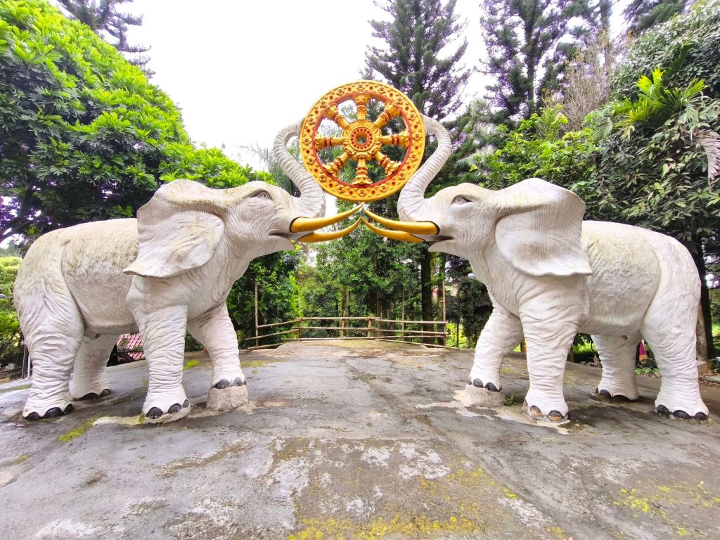 Two stone elephant statues facing each other, trunks raised to touch a gold ornamental wheel between them, in a lush garden setting.