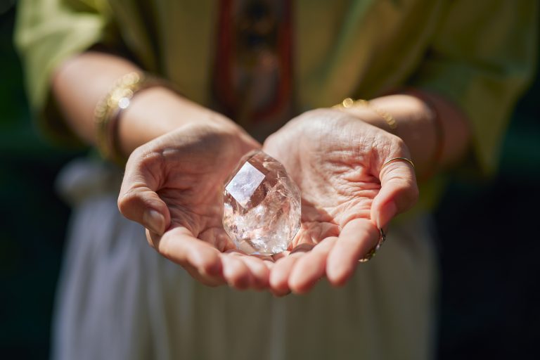 Hands cupping a large clear quartz crystal outdoors with a blurred green background.