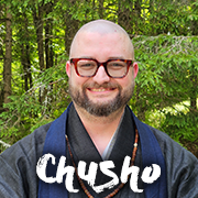 Portrait of a smiling man with red glasses and a beard, outdoors in a forest, with the word 'Chusho' across the bottom.