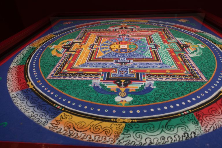 Colorful circular mandala painted with bright geometric patterns in blue, green, red, and yellow on a blue background.